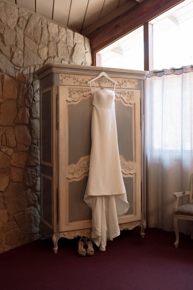 A finished gown hanging on a carved armoire with the bride's shoes on the floor below