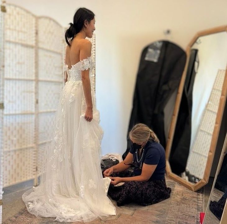Roya kneeling on the studio rug pinning the hem of a lace wedding gown while the bride stands in it, beside a tall wood framed mirror
